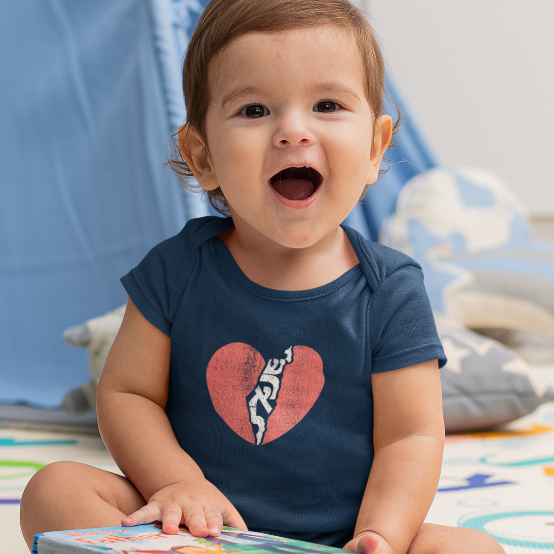 A baby boy in a navy blue onesie with a red heart and the word "Israel" written in Hebrew down the middle.