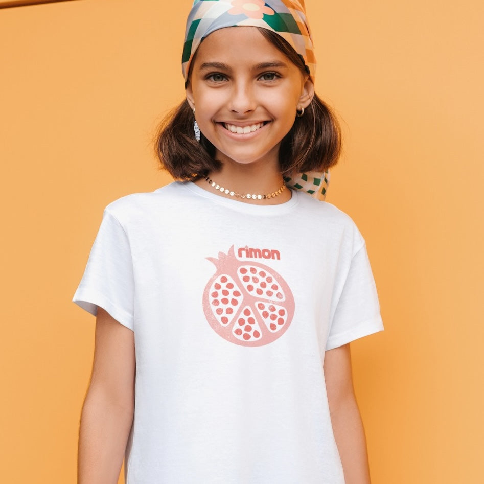 A young girl wearing a bandana on her head and a white t-shirt with an illustration of a pomegranate.