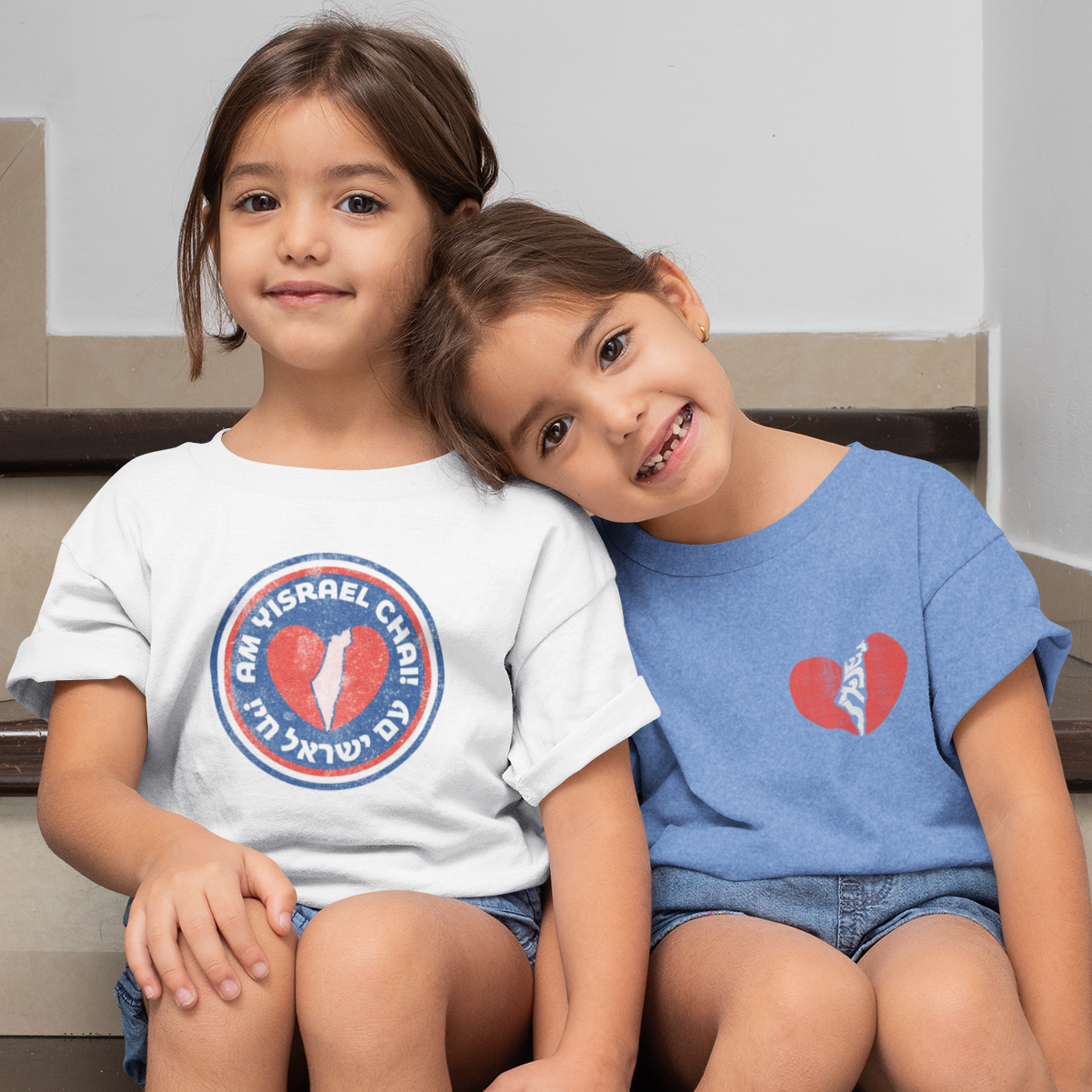 Twin toddler girls sitting together, one in a white t-shirt that reads, "Am Yisrael Chai," the other in a blue t-shirt with a heart and a silhouette of Israel.