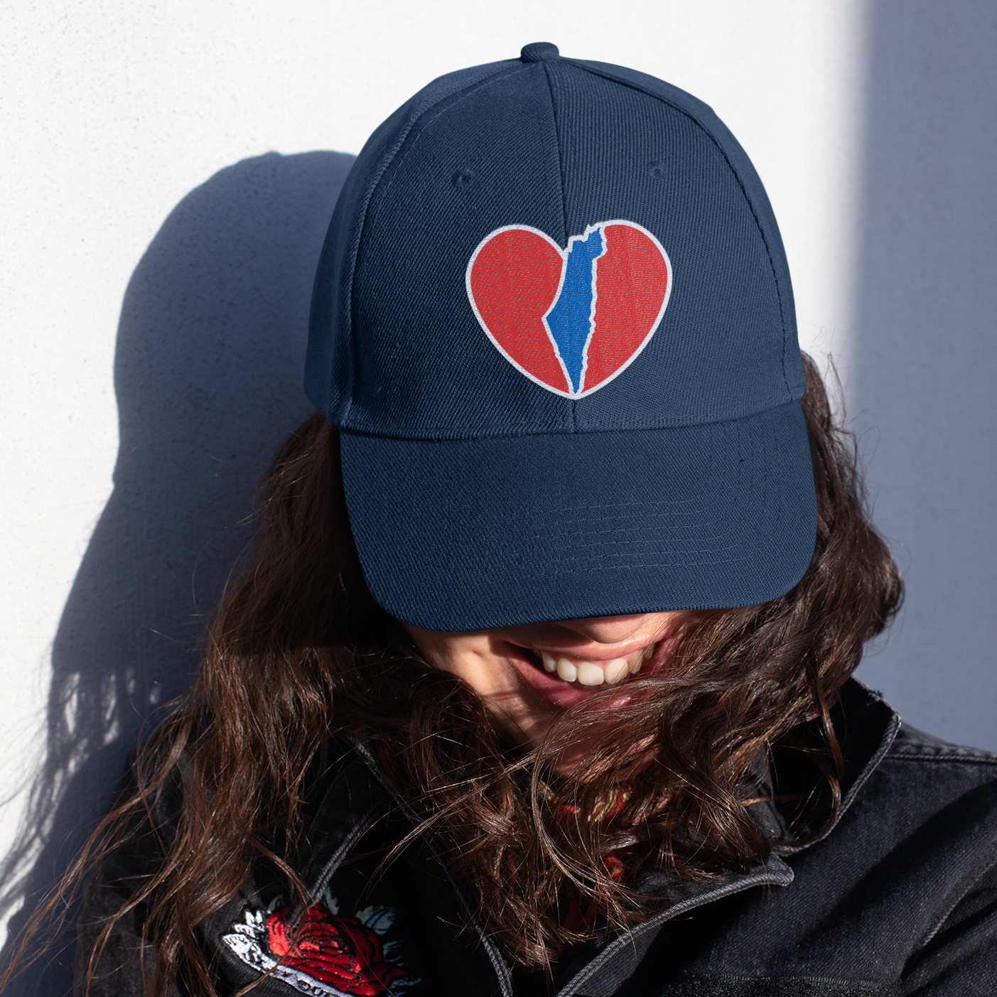 A young woman with her head tiled town, wearing a baseball cap featuring a heart with the shape of Israel in the center.